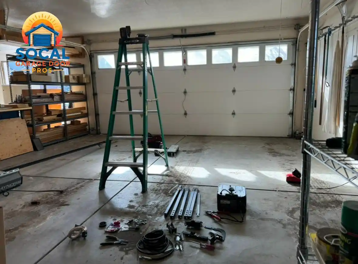 Interior of a garage with a closed white sectional door featuring a row of rectangular windows. A tall green ladder stands in the center, while metal rails, cables, hand tools, hardware, and equipment are spread across the concrete floor beside storage shelves.