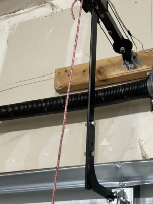 Close-up of a black garage door torsion spring mounted on a horizontal shaft above the door, with a central support bracket and opener arm attached to wooden backing. A red-and-white emergency-release cord hangs in front of the unfinished wall.