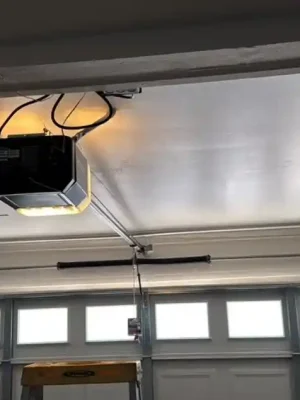 Interior view of a closed gray sectional garage door with a row of rectangular windows, metal tracks, and a horizontal torsion spring mounted above it. A ceiling-mounted garage door opener is illuminated, with wiring and a hanging release cord visible overhead.