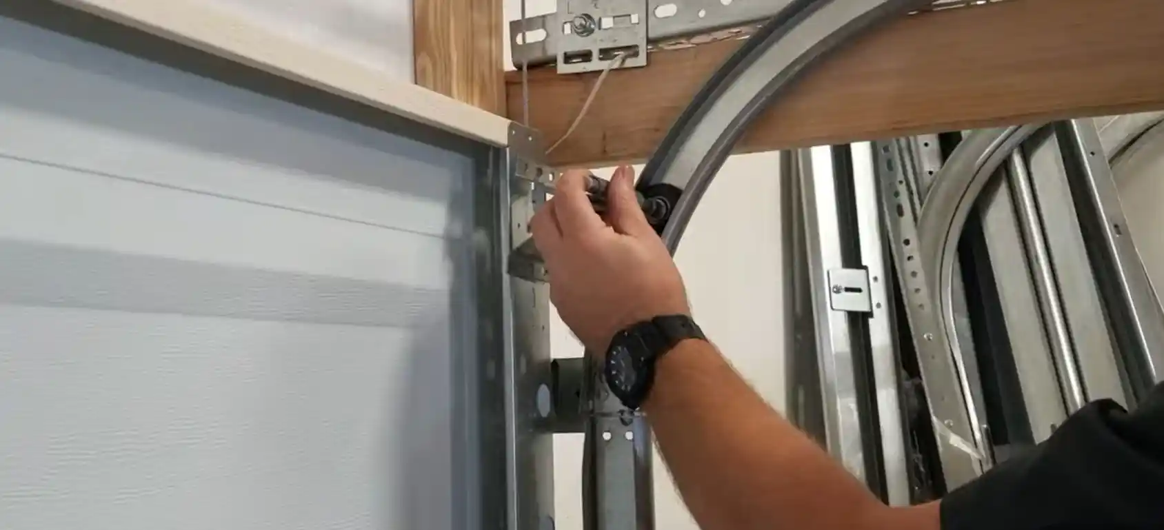 Close-up of a person holding a black garage door roller near the upper corner of a sectional door, beside the curved metal track, support brackets, and wooden framing.