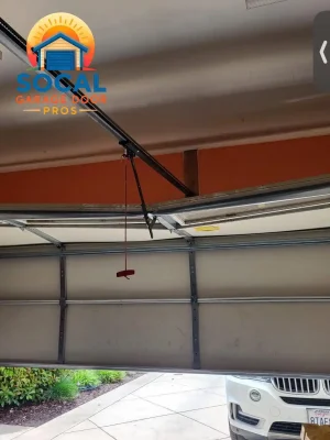 Interior view of a partially open sectional garage door with metal reinforcement rails, hinges, tracks, and a black opener arm connected overhead. A red emergency-release handle hangs near the center, and part of a white SUV is visible outside beside a landscaped driveway.