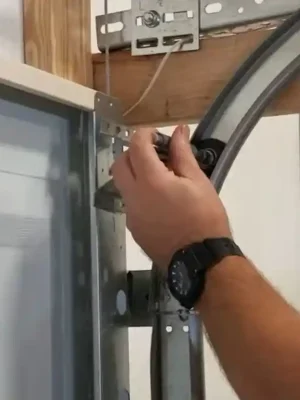Close-up of a person holding a black garage door roller near the upper corner of a sectional door, beside the curved metal track, support brackets, and wooden framing.