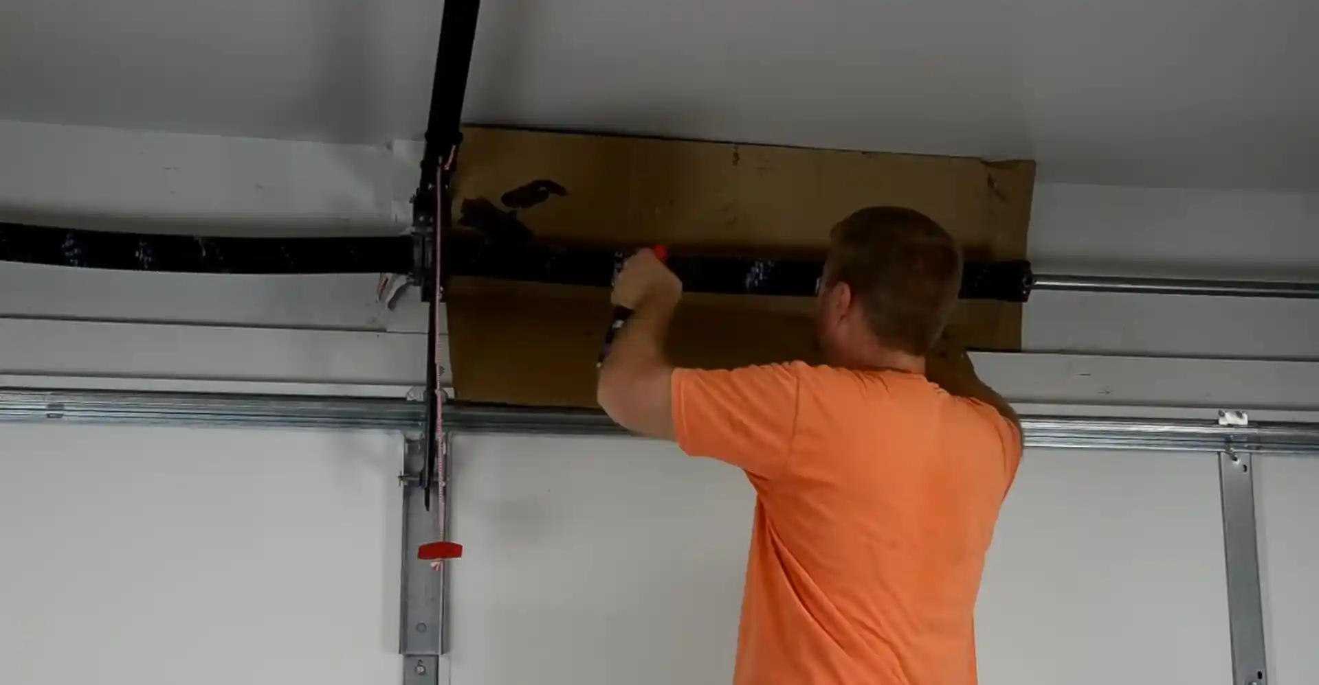 A person in an orange shirt uses a hand tool on the torsion spring assembly mounted above a white sectional garage door. A cardboard panel is positioned behind the spring and metal shaft.