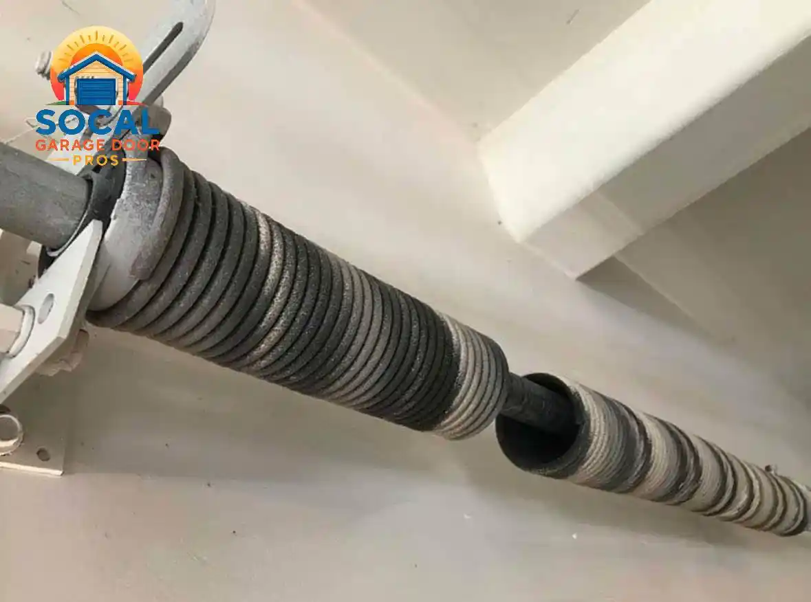 Close-up of a garage door torsion spring assembly mounted near a white ceiling, showing a large gap where the dark metal spring has broken around the horizontal shaft.