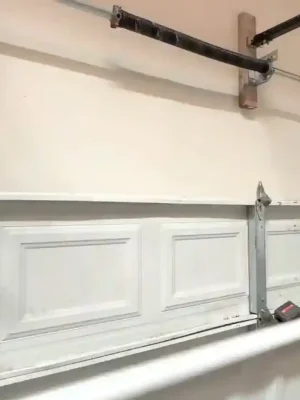 A worker wearing gloves and a black cap lifts a large white replacement panel into position beneath an existing sectional garage door panel. Metal tracks, hinges, reinforcement bars, and the torsion spring assembly are visible around the door.