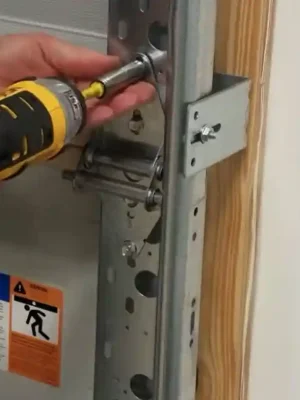 Close-up view of a technician using a yellow and black power drill to tighten a bolt on a metal bracket attached to a garage door track; the bracket is secured to a wooden frame, showing detailed hardware alignment and safety labels on the door panel.