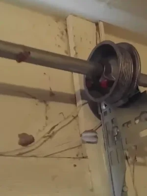 Close-up of a grooved metal garage door cable drum mounted on a horizontal shaft and support bracket near the curved door track. A thin steel cable runs from the drum along the pale wooden wall.