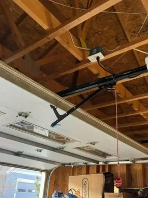 Interior of an unfinished wooden garage with an open white sectional door, exposed ceiling beams, metal tracks, and a ceiling-mounted opener. Cardboard boxes and stored items sit along the wall, while daylight enters through the open doorway.