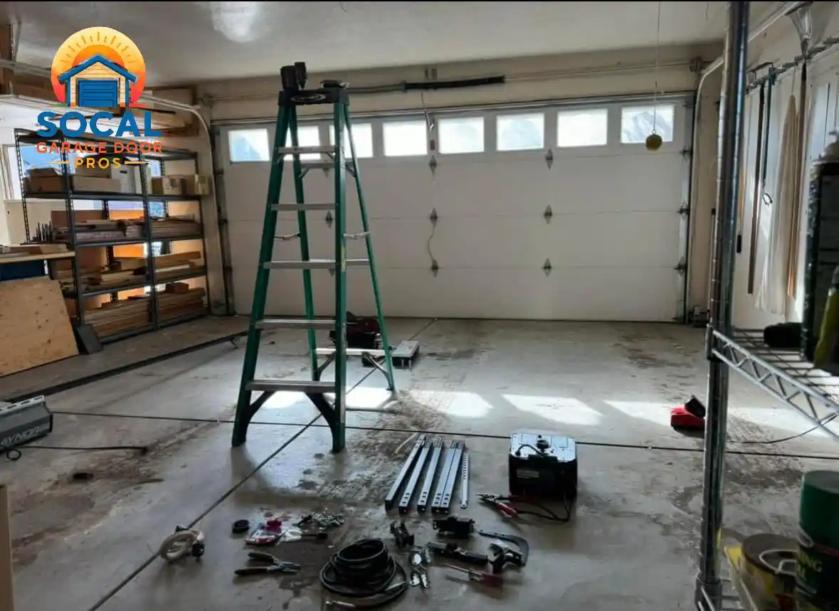 Interior of a garage with a closed white sectional door featuring a row of rectangular windows. A tall green ladder stands in the center, while metal rails, cables, hand tools, hardware, and equipment are spread across the concrete floor beside storage shelves.