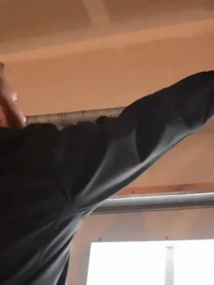 A person in a dark jacket sprays lubricant from an aerosol can onto a garage door torsion spring and metal hardware mounted above the door.