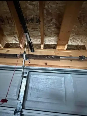 Interior close-up of the upper section of a white garage door with a horizontal torsion shaft, center mounting bracket, opener rail, and metal support arm. Exposed wooden ceiling beams and plywood sheathing are visible above, with a red emergency-release handle hanging down.
