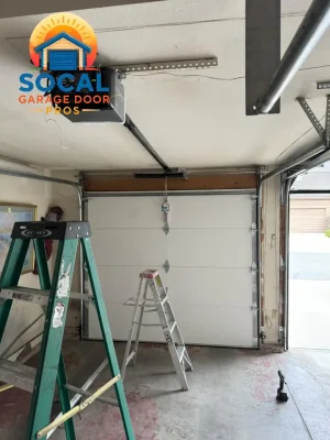 Interior of a garage with a closed white sectional door, exposed metal tracks, and a ceiling-mounted opener connected by a black rail. A large green ladder and a smaller aluminum step ladder stand on the worn concrete floor, with a cordless drill near the open doorway.