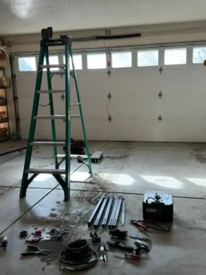 Interior of a garage with a closed white sectional door featuring a row of rectangular windows. A tall green ladder stands in the center, while metal rails, cables, hand tools, hardware, and equipment are spread across the concrete floor beside storage shelves.
