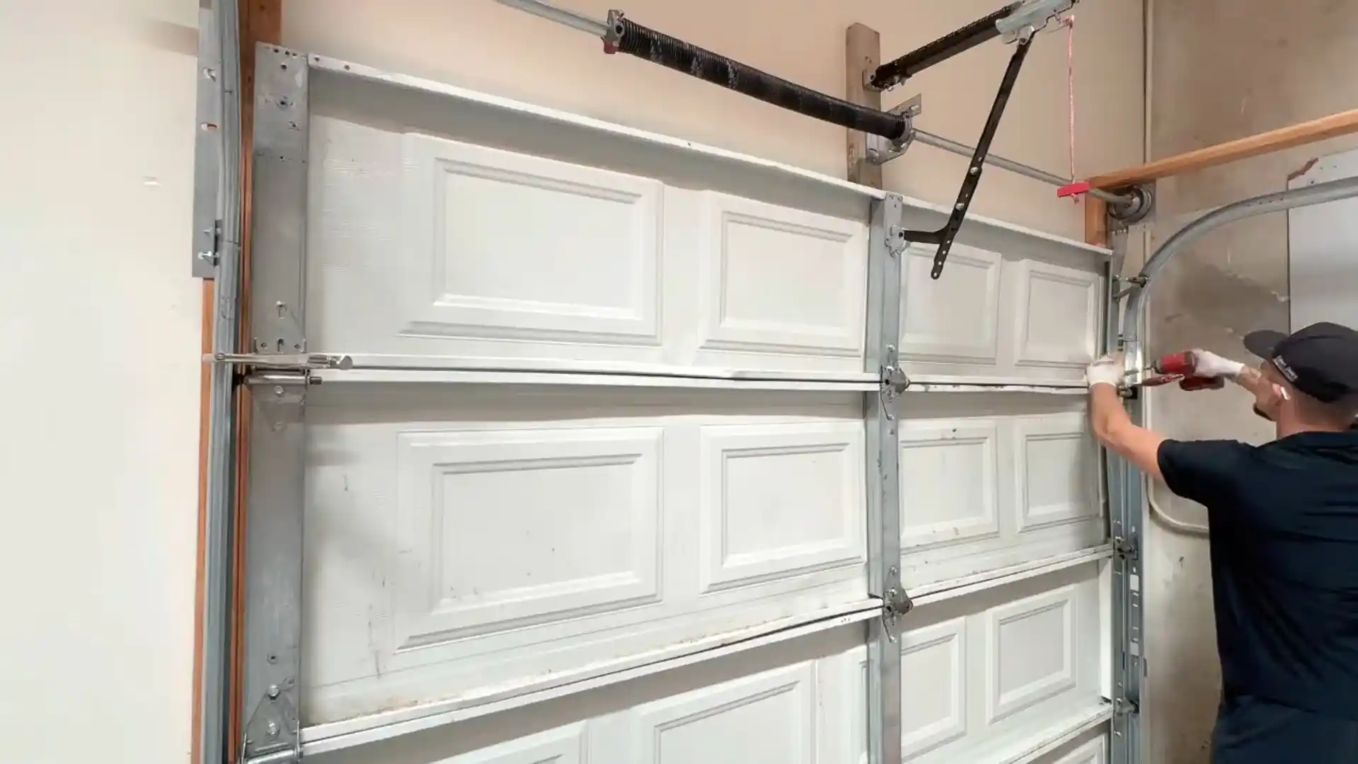 A worker wearing gloves uses a red power drill on the right-side hinge and track of a white sectional garage door. Metal reinforcement bars, rollers, curved tracks, torsion springs, and opener hardware are visible around the door.